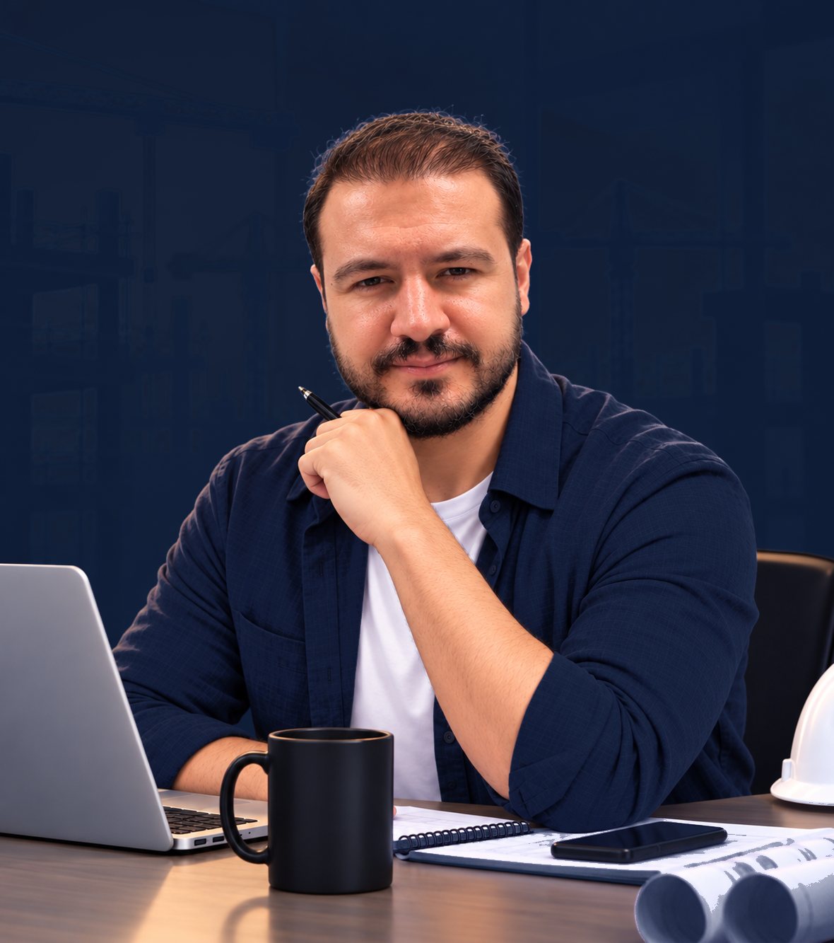 Barış Emre Özçelik at a desk with drawings and a laptop, a site behind